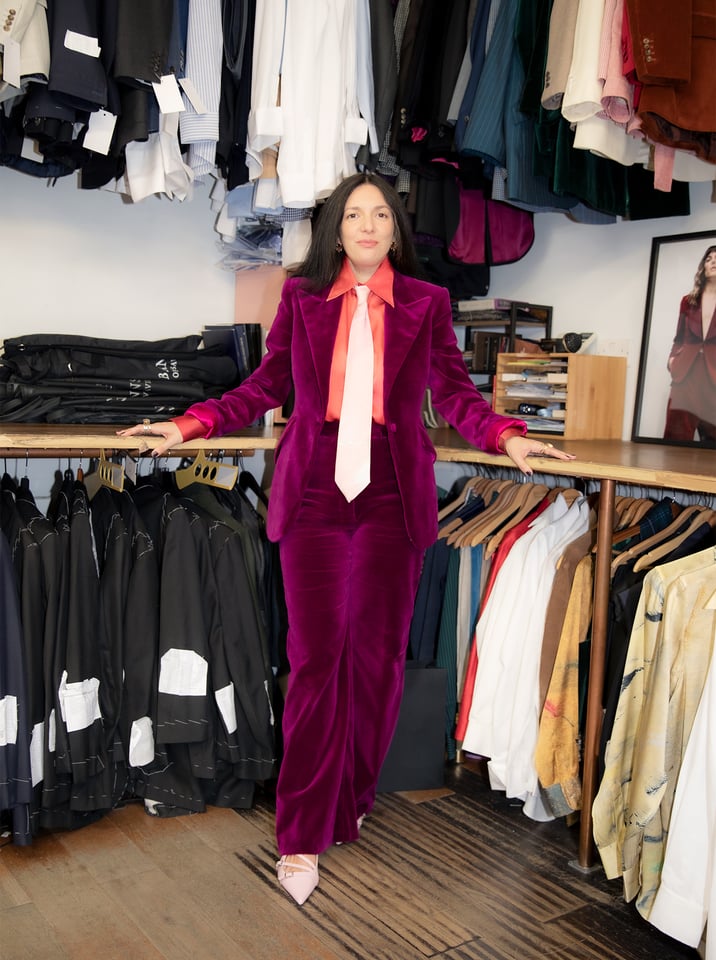 A woman in a bold magenta velvet suit poses confidently in a tailored clothing shop, surrounded by hanging garments.